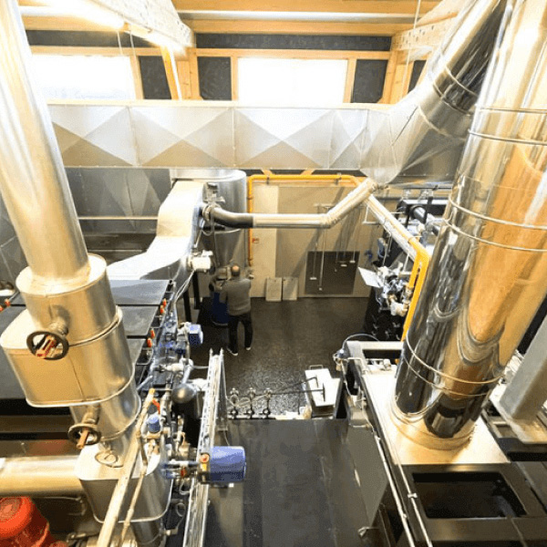 Eine Person steht in einer großen Industrieanlage im Aurora Tierkrematorium, umgeben von Metallrohren, Maschinen und Lüftungsschächten bei heller Beleuchtung. Aurora Tierkrematorium