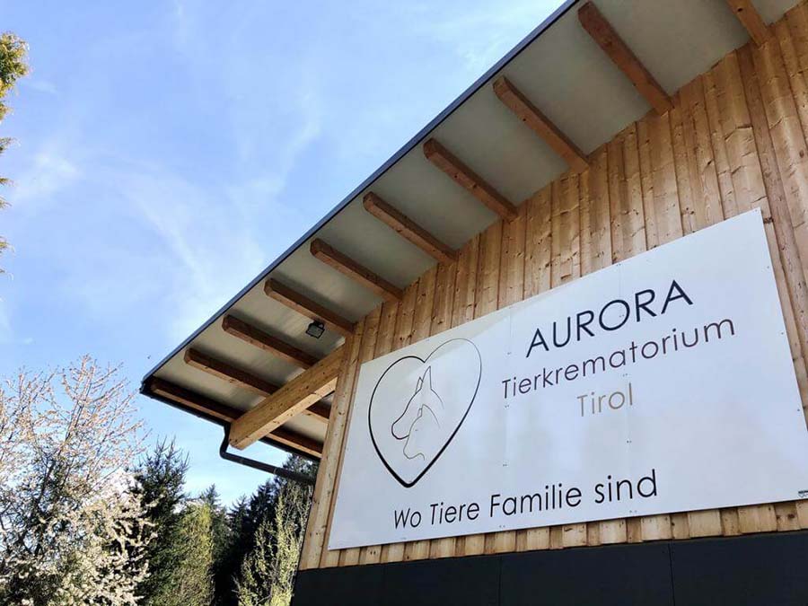 Holzgebäude mit einem Schild mit der Aufschrift "AURORA Tierkrematorium Tirol" und "Wo Tiere Familie sind", mit Bäumen und blauem Himmel im Hintergrund. Aurora Tierkrematorium
