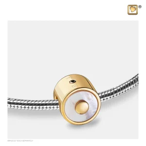 Eine Nahaufnahme eines goldenen und weißen kreisförmigen Anhängers mit einer perlenartigen Mitte an einem silbernen Armband. Das Armband ist separat mit einem Markenlogo in der oberen rechten Ecke abgebildet. Aurora Tierkrematorium