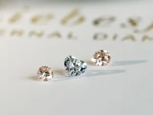 Drei lose Diamanten, zwei im Rundschliff und einer in Herzform, sind auf einer weißen Fläche mit verschwommenem Goldtext im Hintergrund platziert. Aurora Tierkrematorium