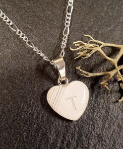 Eine silberne Halskette mit einem herzförmigen weißen Anhänger mit einfacher Liniengravur, der auf einer dunklen Fläche neben einem getrockneten Zweig angebracht ist. Aurora Tierkrematorium