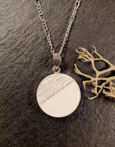 Silberne Halskette mit einem runden weißen Anhänger mit diagonalen schwarzen Linien und einer Reihe kleiner Punkte, die auf einer dunklen, strukturierten Oberfläche neben einem Zweig angebracht sind. Aurora Tierkrematorium