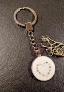 Ein silberner Schlüsselanhänger mit einem runden weißen Anhänger mit kleinen Steinen, die in Herzform angeordnet sind, auf einer dunklen Oberfläche neben einer getrockneten Pflanze. Aurora Tierkrematorium