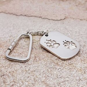 Ein silberner Schlüsselanhänger mit Karabinerhaken und einem rechteckigen Anhänger, auf dem zwei Pfotenabdrücke eingraviert sind, ruht auf einer strukturierten Steinoberfläche. Aurora Tierkrematorium